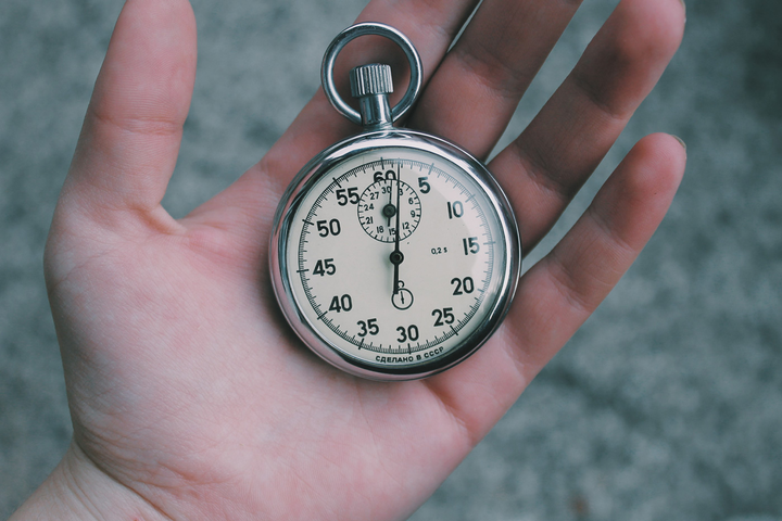 Silver stopwatch held in an open hand with the dial facing the camera, showing about 0–5 seconds elapsed on a white clock face.