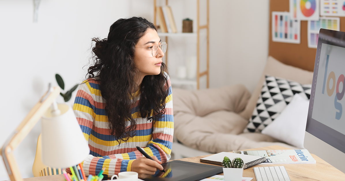Woman with curly hair and glasses sits at a desk, using a graphics tablet while looking at a computer screen in a bright, colorful workspace.
