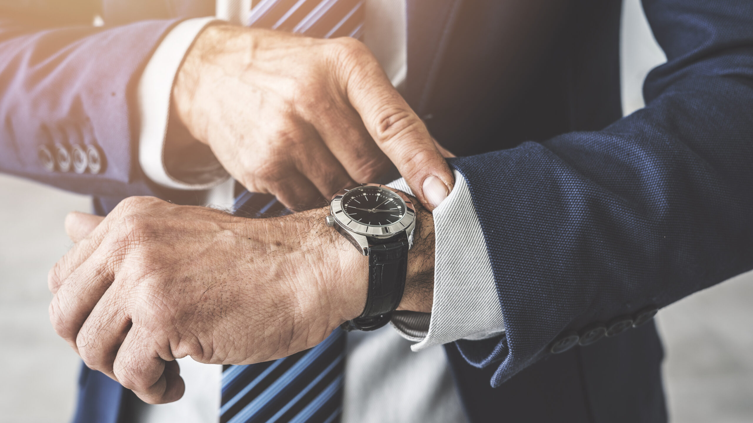 Close-up of a man in a navy suit adjusting a silver wristwatch on his left wrist.