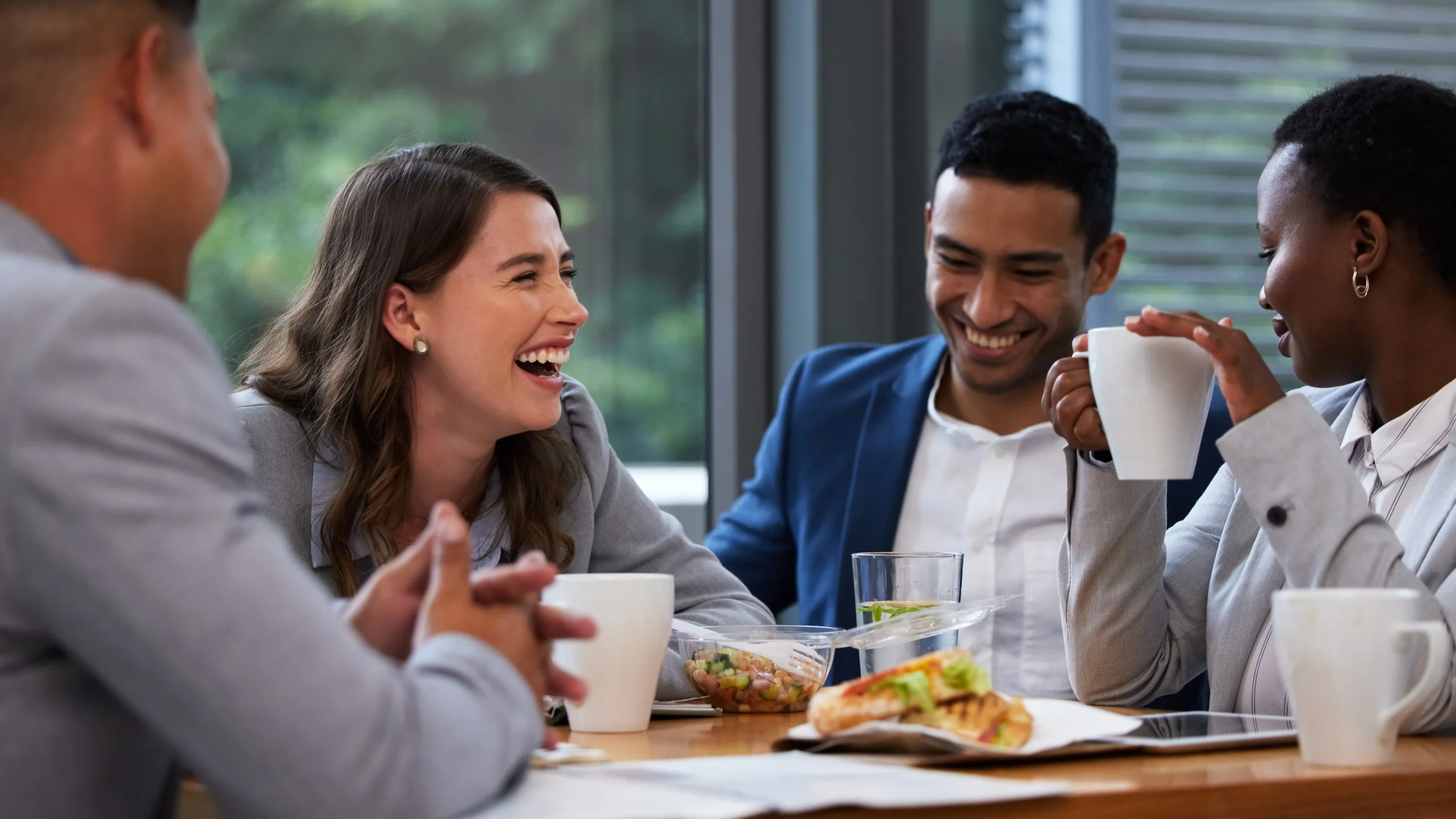 Group of diverse coworkers laughing and talking at a table with coffee mugs and lean sandwiches in a bright, casual meeting setting.