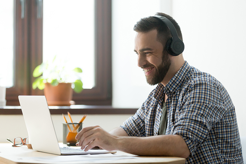 Man wearing headphones, typing on a laptop at a bright desk in a home office, smiling while working.