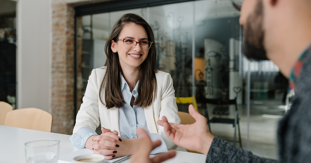Smiling woman with dark hair, red glasses, and a white blazer sits at a table as a colleague talks and gestures nearby in a modern office.