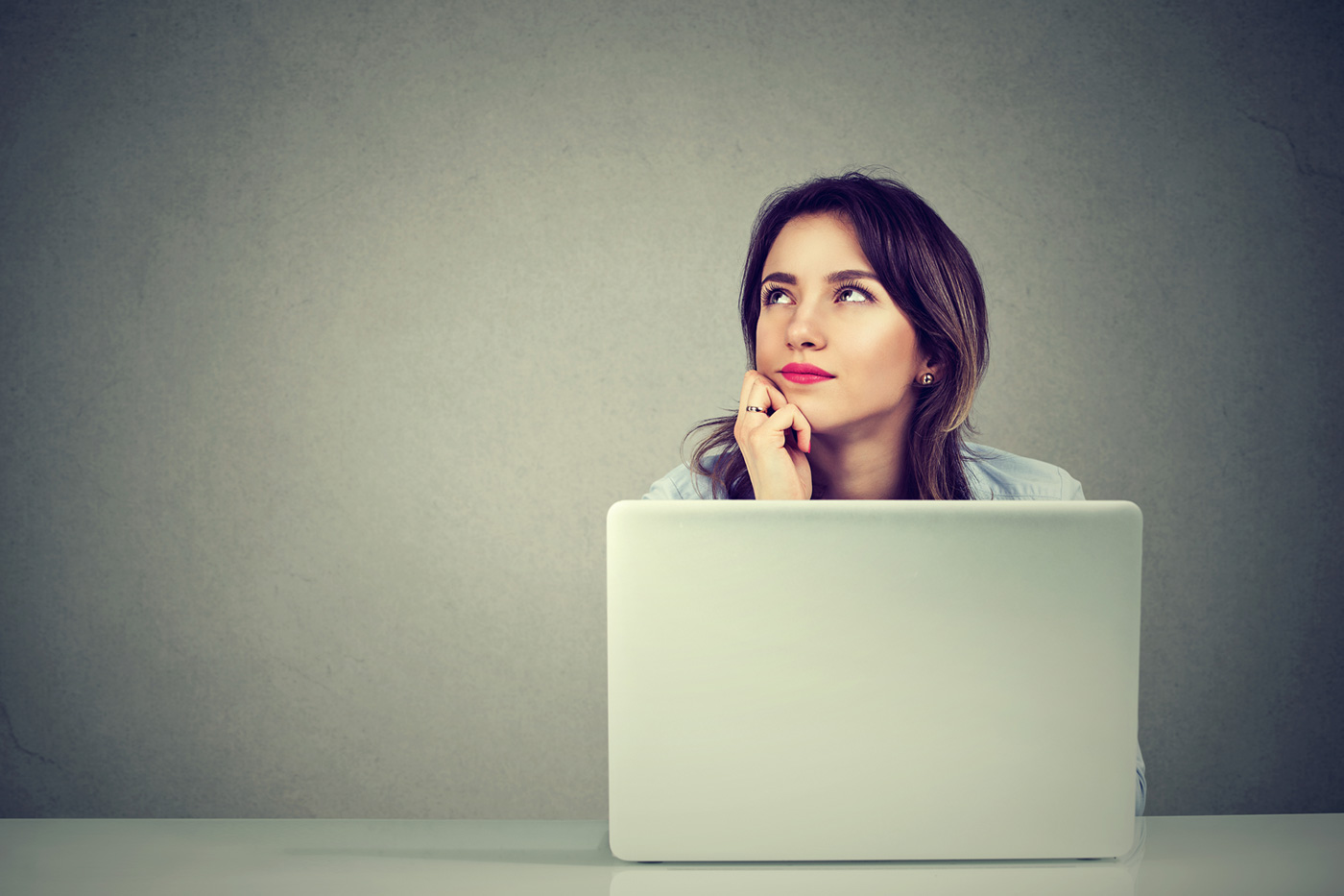 Woman sitting at a desk with a laptop, chin resting on her hand, looking upward in thought.