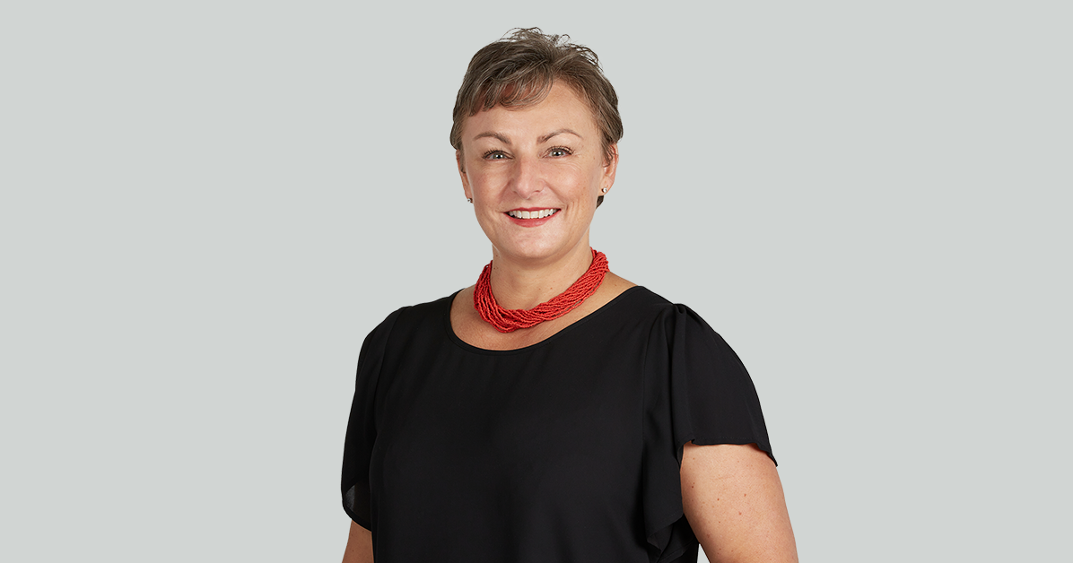 Professional portrait of a smiling woman wearing a black blouse and red necklace against a light gray background.