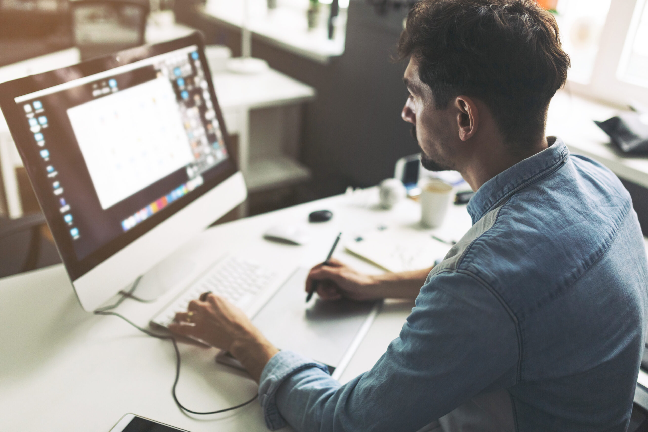Man in denim shirt sits at a desk, working on a desktop computer with a pen in hand and a tablet nearby in a bright office.
