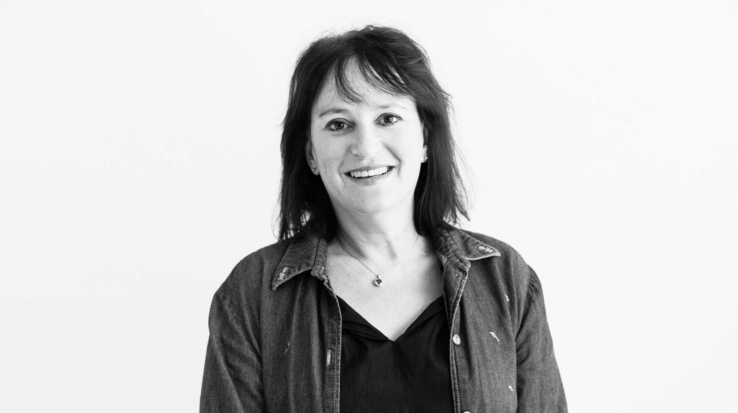 Portrait of a smiling woman with dark hair wearing a denim jacket against a light background, facing the camera.