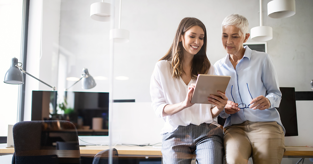 Two women sit together at a desk in a bright office, smiling while looking at a tablet screen together—collaborative work.