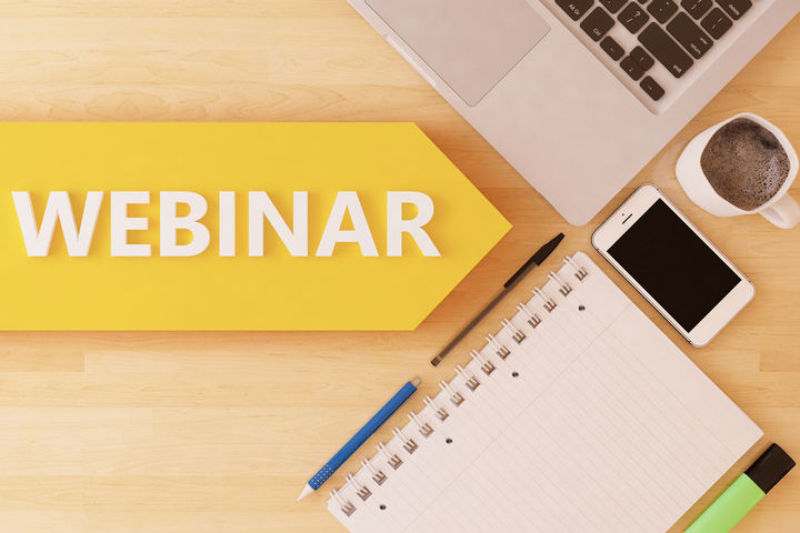 Yellow arrow sign spelling WEBINAR on a desk with a laptop, notebook, phone, pencil, and coffee cup.