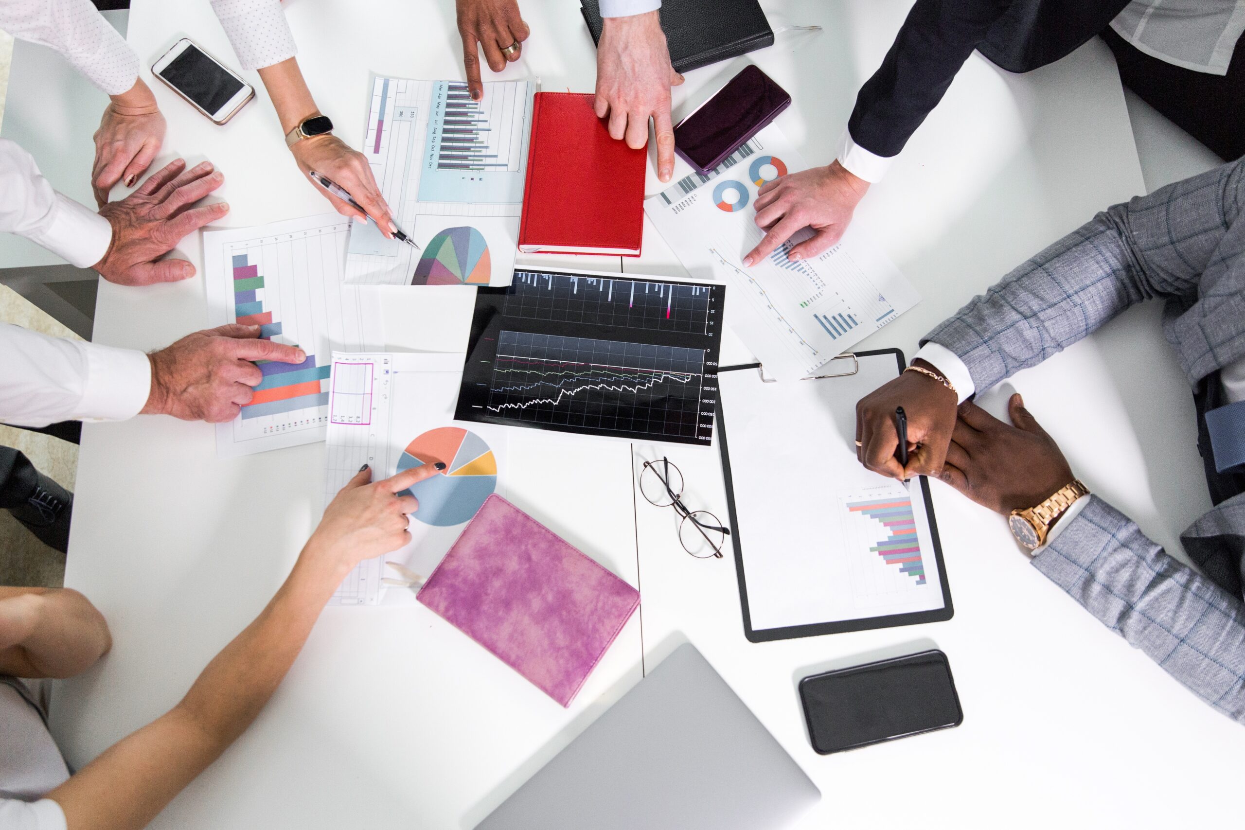Top-down view of professionals around a table reviewing charts and graphs with tablets and notebooks.