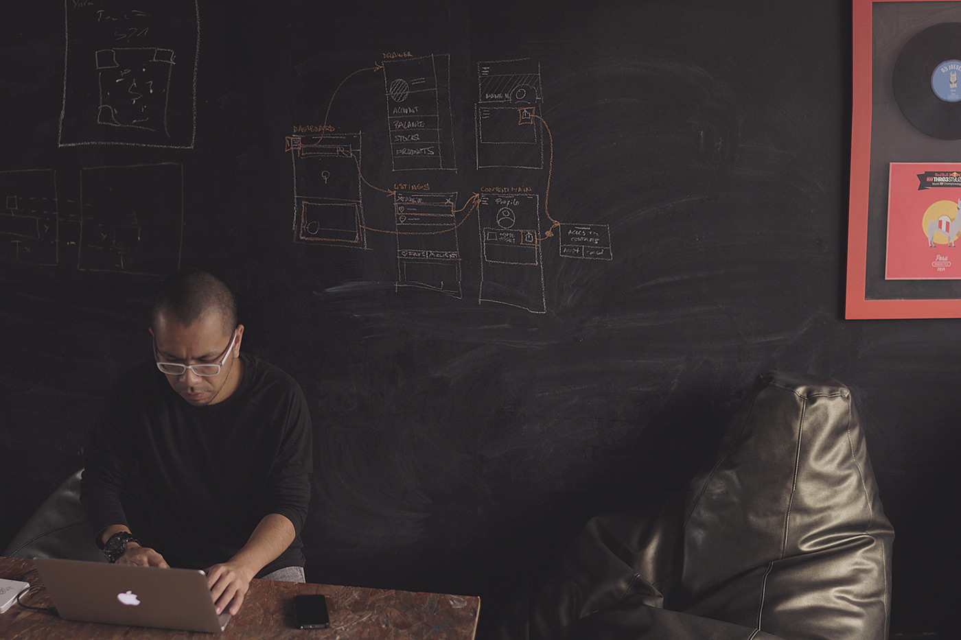 Man with glasses typing on a silver laptop at a wooden table in a room with a chalkboard wall and a beanbag chair to the right.