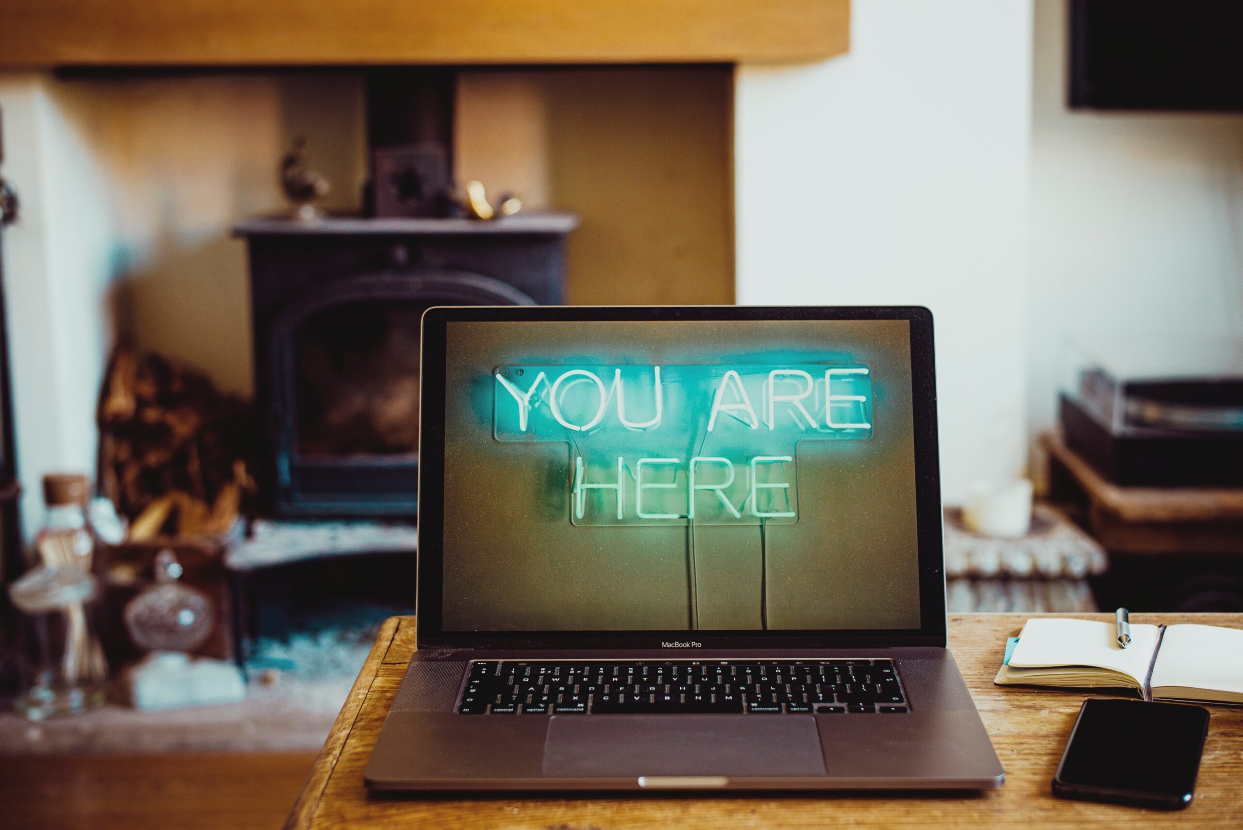 Laptop on a wooden desk showing a neon 'YOU ARE HERE' sign on the screen, with a blurred living room and fireplace in the background.