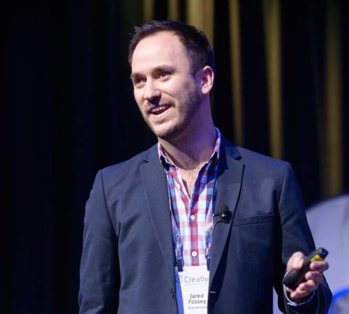 Man in a dark blazer giving a talk on stage, wearing a plaid shirt and a conference badge, holding a presentation remote with a microphone clipped to his lapel.