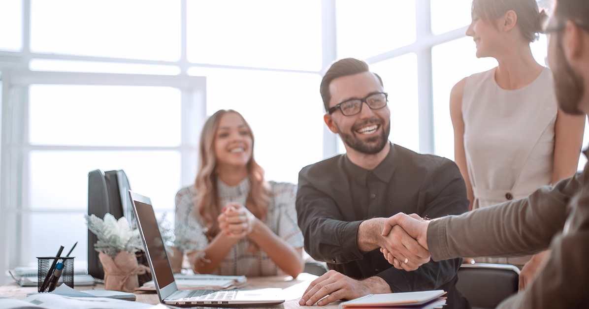Business colleagues celebrate an agreement with a handshake across a desk in a bright office, smiles all around.