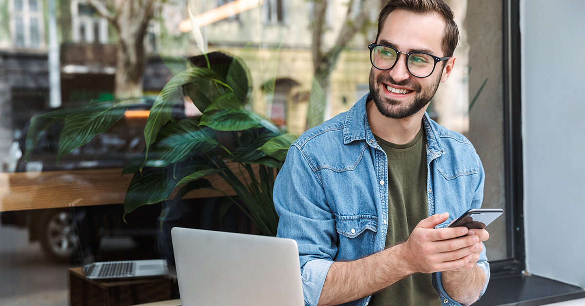 Smiling man with glasses in a denim shirt at a cafe, looking at his phone with a laptop nearby.