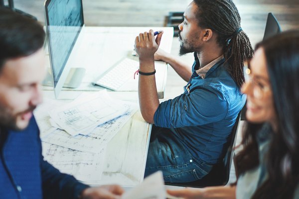 Three coworkers collaborate at a bright desk, reviewing blueprints and a computer screen.