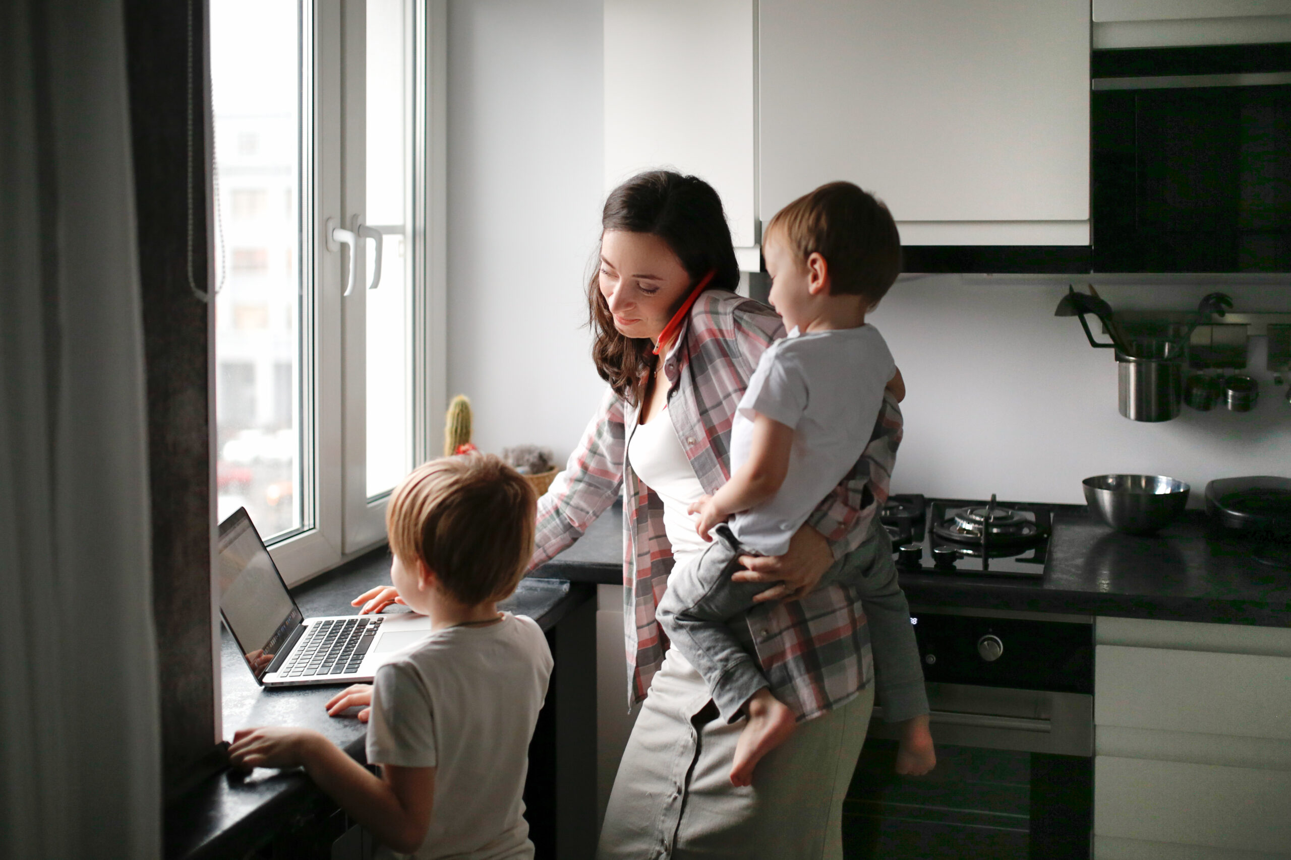 Mother helping two children with a laptop at a kitchen counter by a bright window.