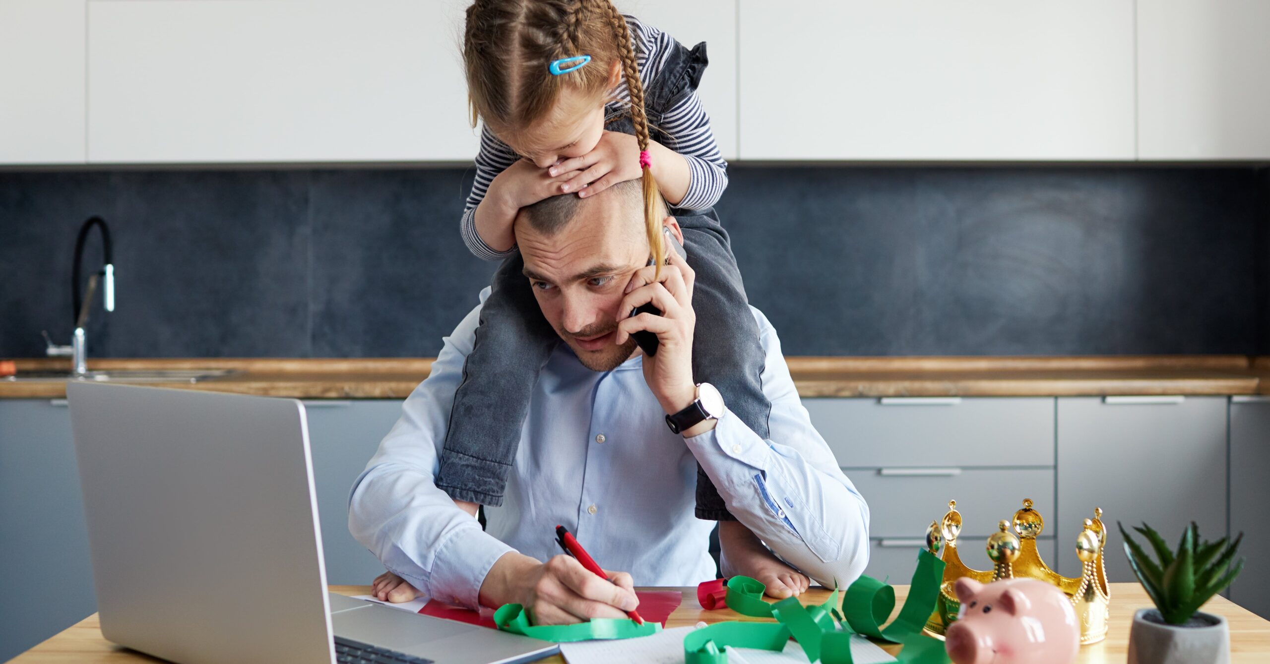 Father with a young girl on his shoulders, cutting colorful paper at a desk with a laptop nearby.