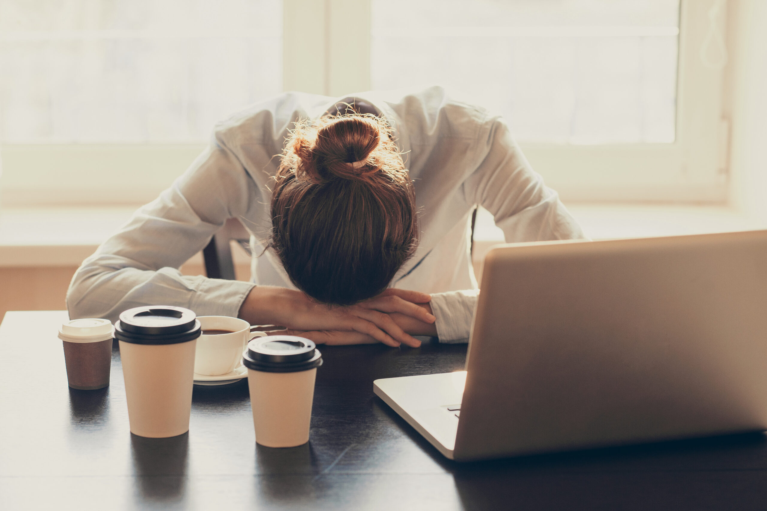 Person with a bun rests their head on folded arms at a desk, in front of a laptop and several takeaway coffee cups.