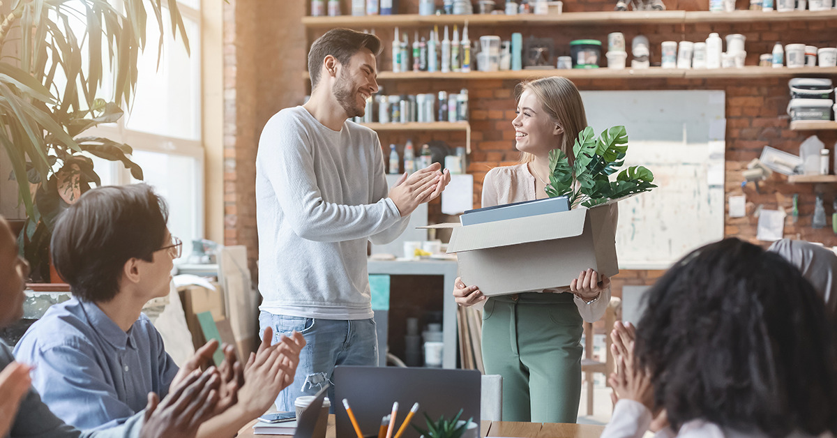 Colleagues cheer as a woman carries a cardboard box with a plant and blue folders into a bright, casual office space.