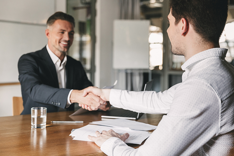 Two businessmen shake hands across a desk, with papers and a glass of water on the table.