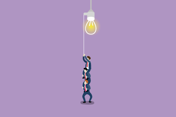Three people in business attire stack on a rope to lift a glowing light bulb, symbolizing teamwork.