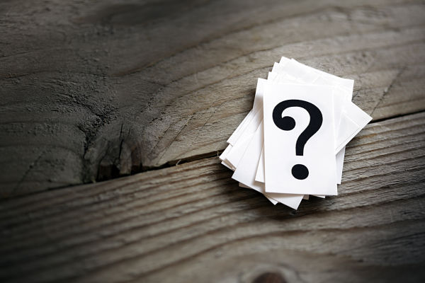 Stack of white cards with a large black question mark on a weathered wooden surface, symbolizing a question or unknowns.