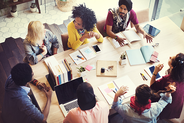 Eight people of diverse backgrounds gathered around a light wood table, collaborating with laptops, notebooks, and tablets in a bright coworking space.