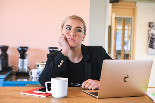 A woman in a black blazer sits at a desk with a laptop, resting her chin on her hand in a thoughtful pose, coffee mug and notebook nearby.