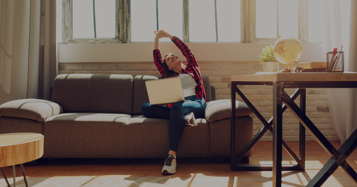 Woman on a sofa stretching with a laptop on her lap in a bright living room.