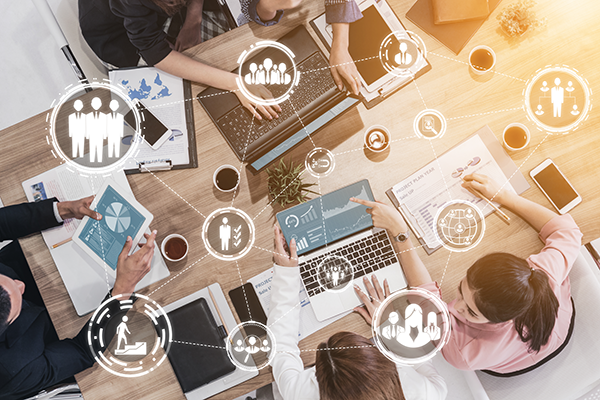 Overhead view of a business team collaborating around a table with laptops, charts, and coffee mugs, surrounded by floating collaboration icons.