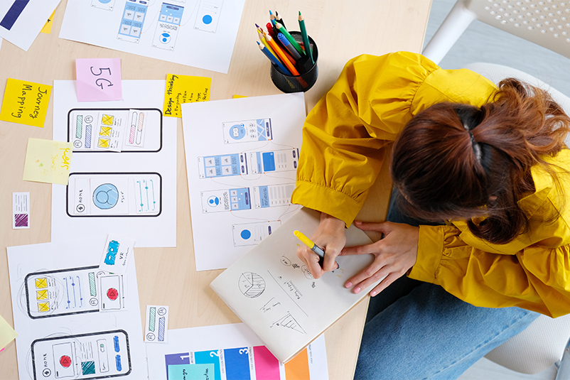 Designer in a yellow jacket writing in a notebook while mobile app wireframes and sticky notes cover the table.