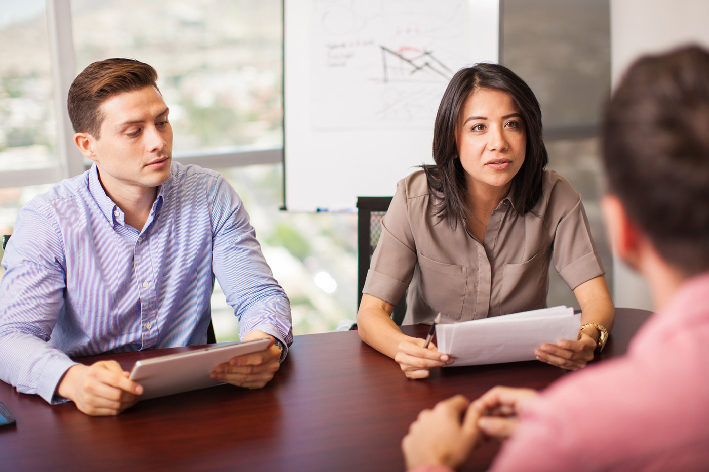 Team meeting around a conference table: woman on the right speaks while holding documents, man on the left looks at a tablet, and a third participant is seen from behind.
