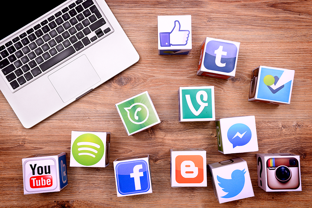 Laptop on a wooden desk surrounded by cube blocks showing social media logos (YouTube, Facebook, Twitter, Instagram, Spotify, Tumblr).