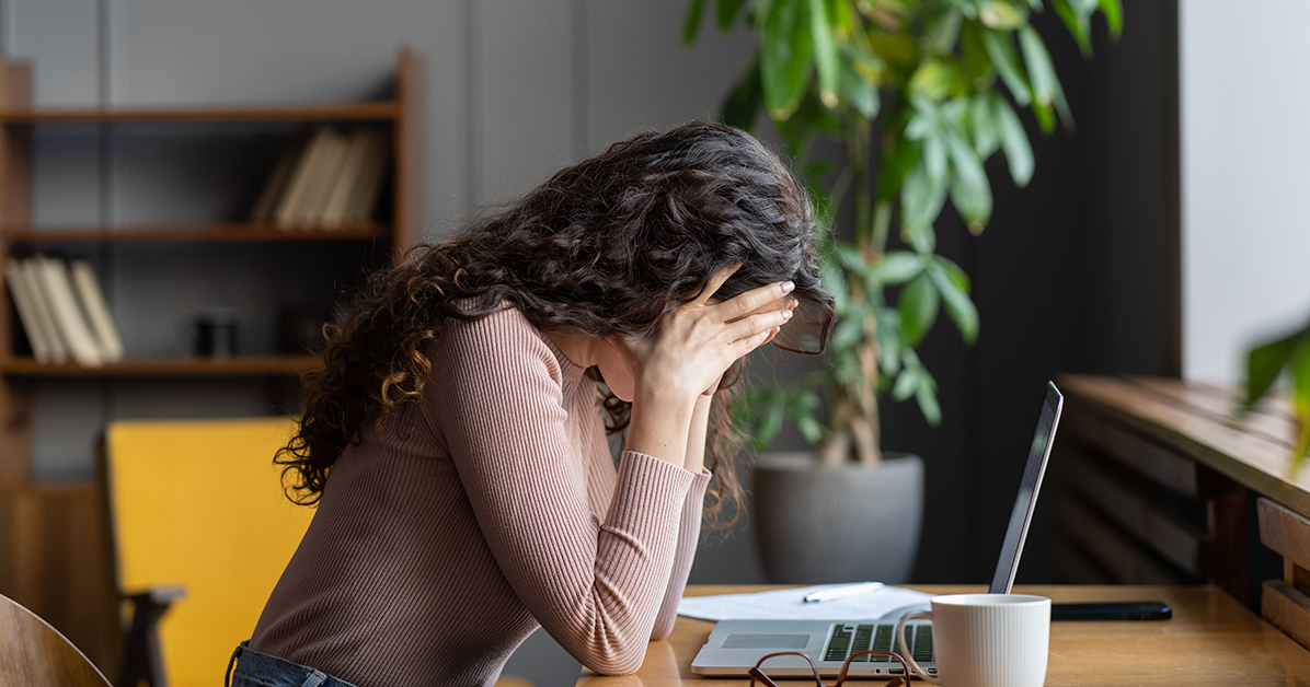 Woman sitting at a desk with her head in her hands, looking stressed while working on a laptop.
