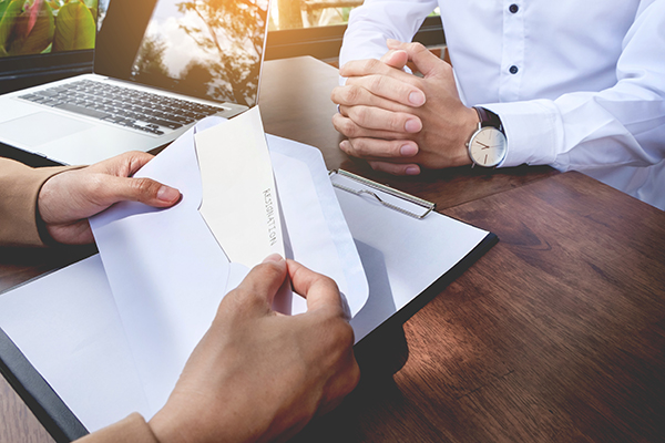 Close-up of a resignation letter being handed over in a business meeting, with a laptop and clipboard on a wooden table.