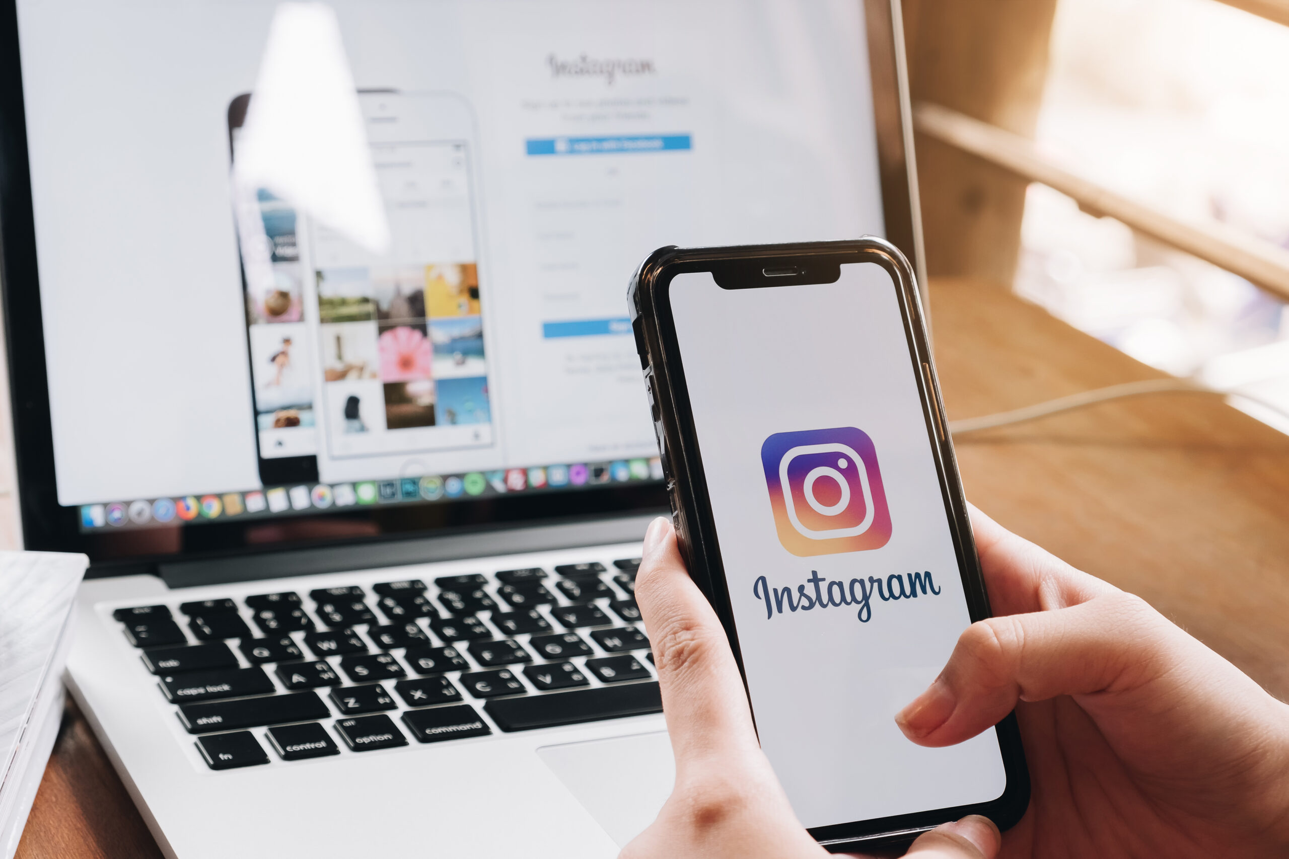 Person holds a smartphone showing the Instagram app icon on screen, with a laptop in the blurred background behind it.