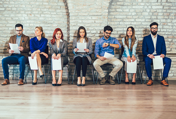 Eight professionals seated in a row against a brick wall, each holding papers and waiting for an interview or meeting.