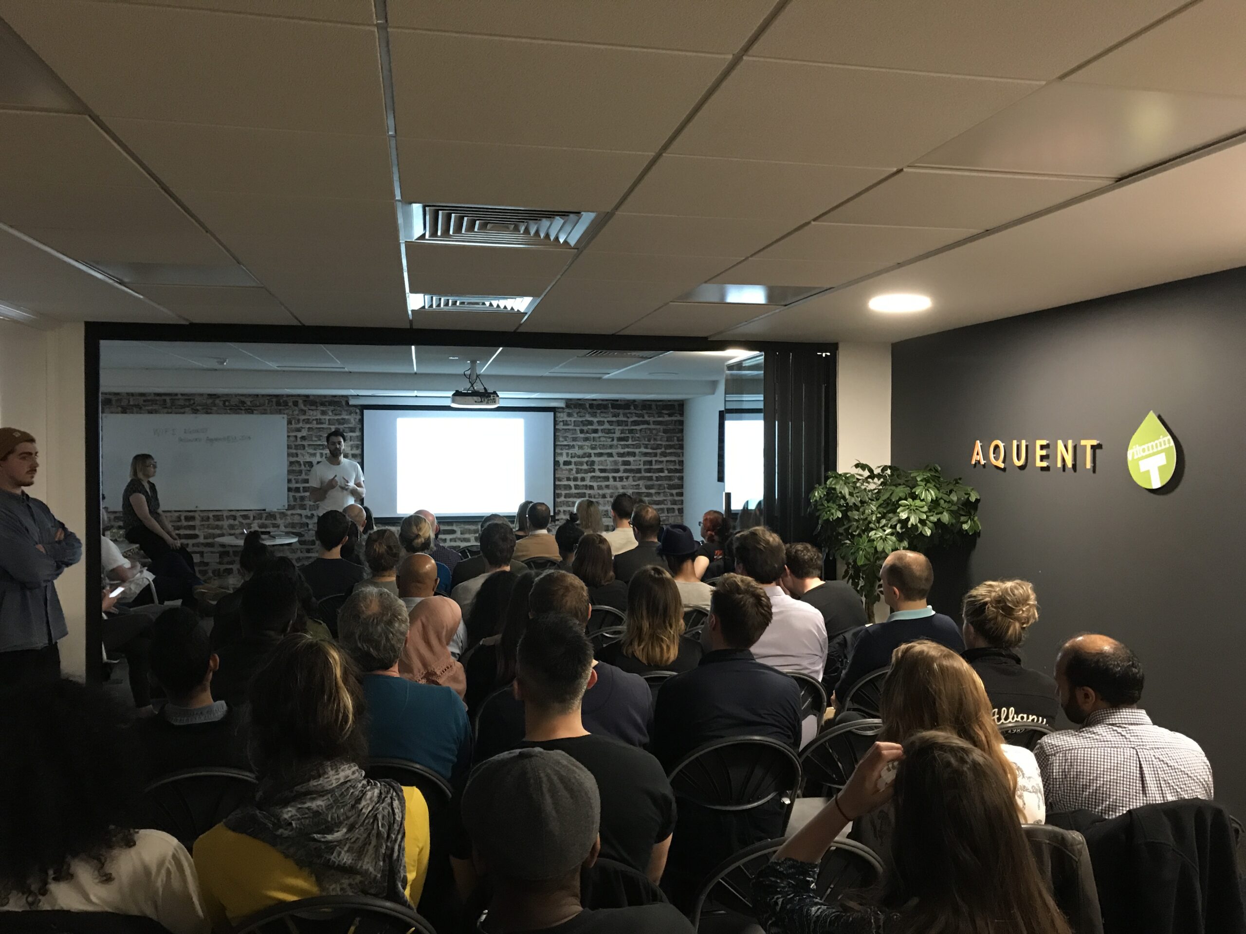 Audience seated in a conference room watching a presenter at the front with a projector screen.