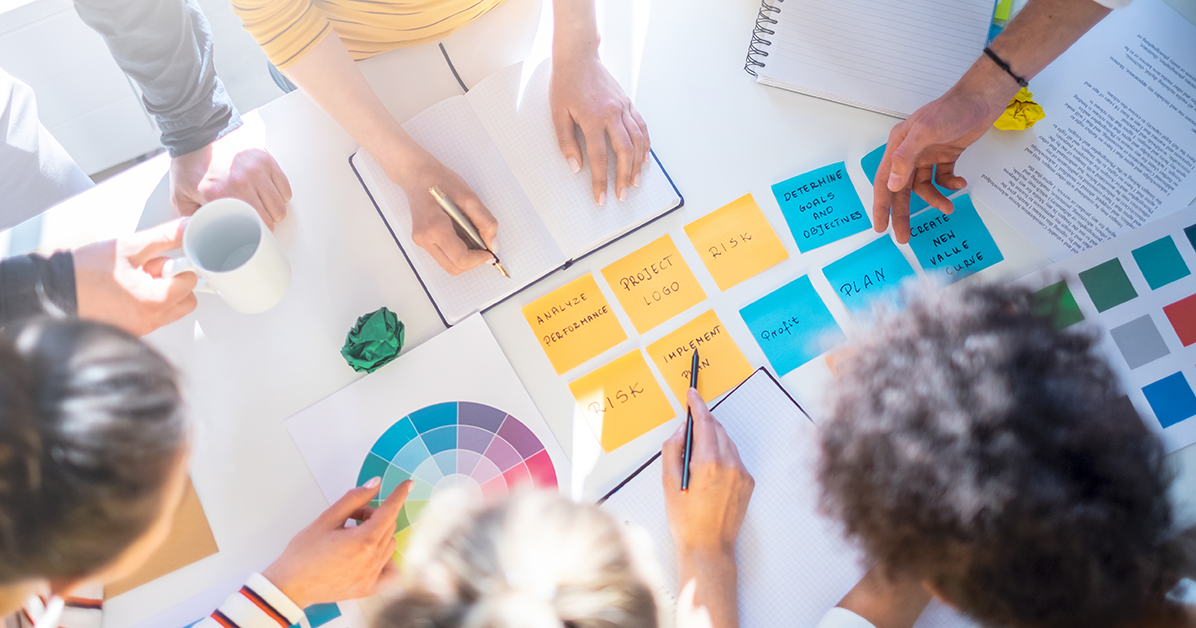 Overhead view of a team brainstorming at a table with sticky notes, notebooks, and color swatches across the surface.