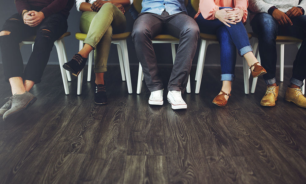 Several people sitting in a row on chairs in a waiting area, showing their legs and shoes.