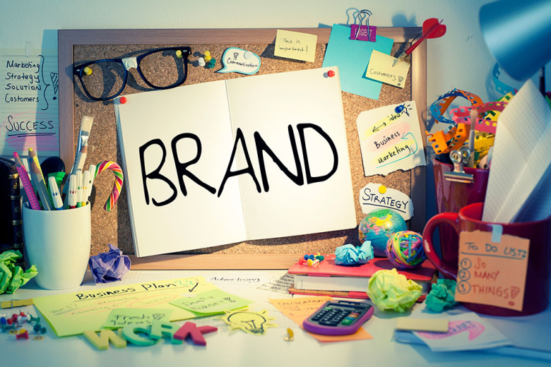 Notebook open to the word BRAND on a corkboard, surrounded by colorful office supplies and sticky notes.
