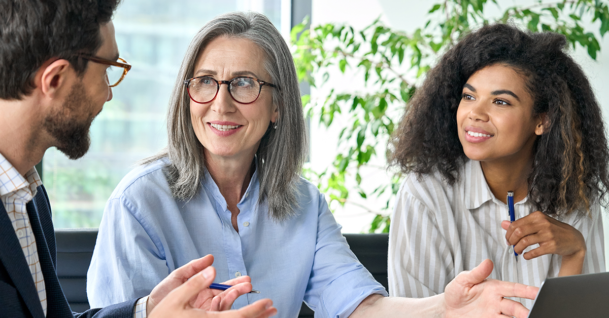 Three coworkers in a bright office discuss a project, the gray-haired woman gesturing with a pen.