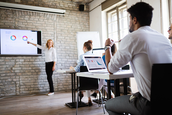 Woman presenter points to a wall screen displaying charts as colleagues watch in a brick-walled meeting room with laptops at the table.