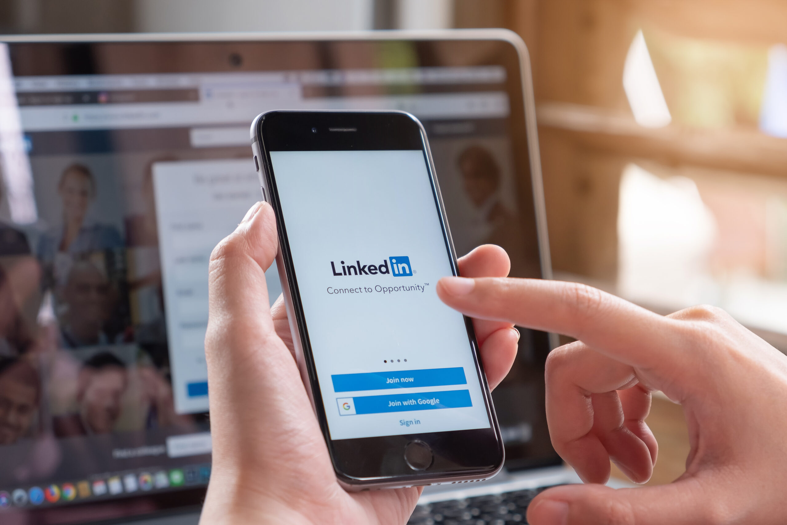 Person holding a smartphone displaying the LinkedIn login screen in front of a laptop monitor, with a blue Join button visible.