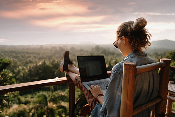 Person with a laptop on a wooden deck overlooking a sunset valley; legs up, wearing sunglasses in a denim jacket.