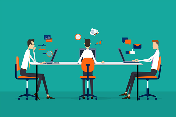 Three coworkers sit on a long table with laptops, collaborating in a modern office.