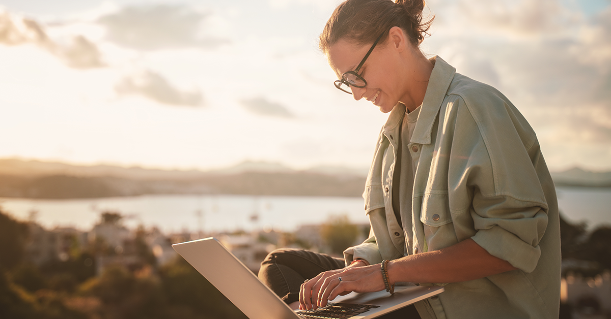 Woman wearing glasses types on a laptop outdoors at sunset with a blurred coastal town in the background.