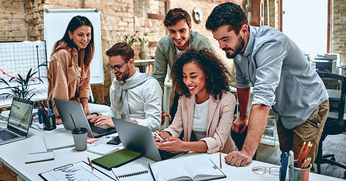Five coworkers gathered around a table, looking at a laptop and tablet while collaborating cheerfully in a bright office space.
