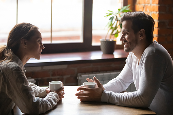 A man and a woman sit across from each other at a wooden table, chatting and holding coffee cups in a sunlit cafe, with a brick wall in the background.