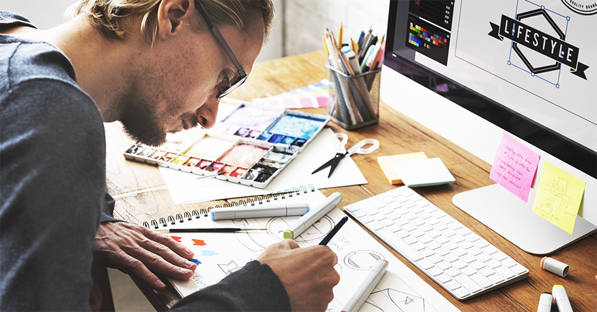 Designer sketching diagrams with markers at a cluttered wooden desk, surrounded by color swatches, tools, and a monitor displaying a lifestyle graphic.